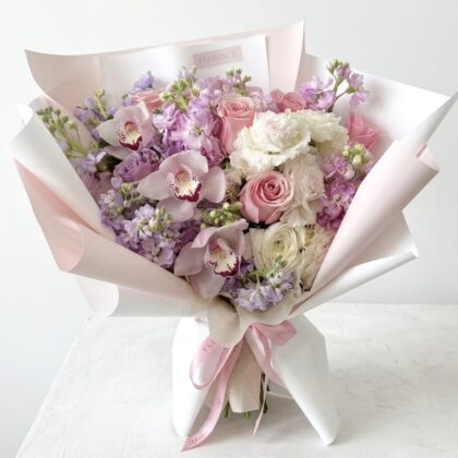 A luxurious bouquet of soft pink roses, lavender stock, and white orchids wrapped in elegant pale pink paper.