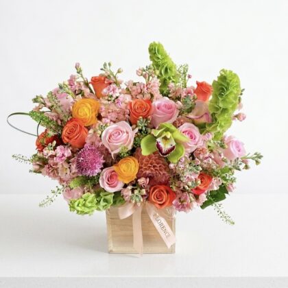 A luxurious floral arrangement of pink and orange roses, green orchids, and bells of Ireland in a wooden box.