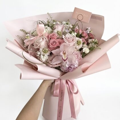 A hand holds a luxurious bouquet of blush pink roses, orchids, and white filler flowers wrapped in elegant pink paper.