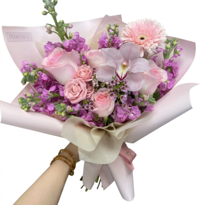 A hand holds a luxurious bouquet of soft pink roses, purple stock, a gerbera daisy, and an elegant cymbidium orchid.