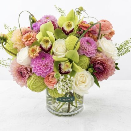 A luxurious floral arrangement of pink dahlias, white roses, and lime green orchids in a clear glass vase.
