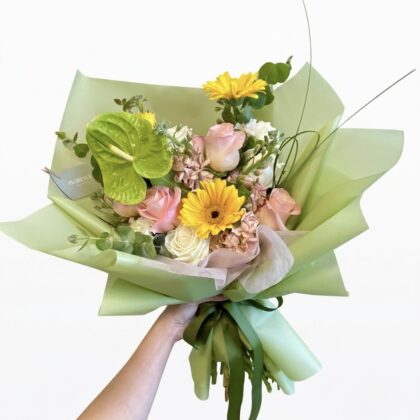 A hand holds a luxury bouquet of yellow gerberas, blush roses, and a green anthurium wrapped in sage paper.