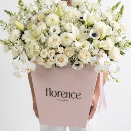 A person holds a large, elegant arrangement of white lilies, roses, and orchids in a blush pink Florence gift box.