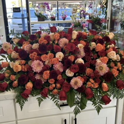 A lush, sprawling casket spray featuring deep red roses, burnt orange roses, and soft pink dahlias with green foliage.