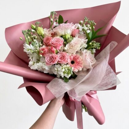 A hand holds a luxurious bouquet of pink and white roses, carnations, and stock flowers wrapped in elegant pink paper.