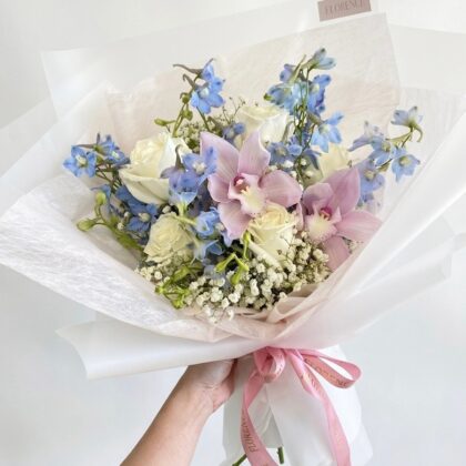 A hand holds a luxury bouquet of white roses, soft pink orchids, and blue delphiniums wrapped in elegant white paper.