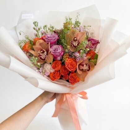 A hand holds a luxurious bouquet of orange roses, lavender roses, and bronze orchids wrapped in elegant white paper.