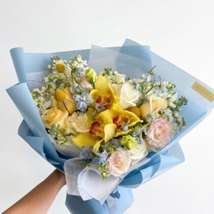 A hand holds a luxurious bouquet of yellow orchids, roses, and blue delphiniums wrapped in elegant light blue paper.