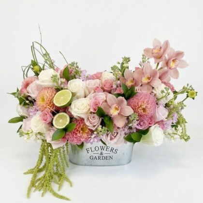 A luxurious floral arrangement of pink dahlias, roses, and orchids in a metal bucket with lime slices and greenery.