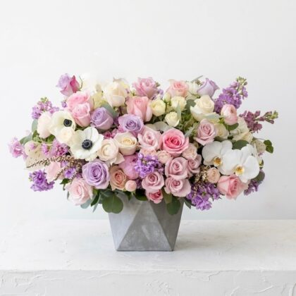 A luxurious floral arrangement of pastel roses, orchids, and anemones in a modern grey geometric concrete vase.