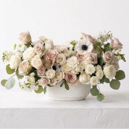 A luxurious floral arrangement of blush roses, white anemones, and eucalyptus in a minimalist white ceramic bowl.
