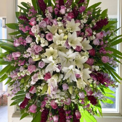 A grand funeral spray featuring white lilies, lavender roses, and deep burgundy snapdragons arranged with lush green palm leaves.