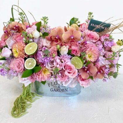 A lush floral arrangement of pink roses, orchids, and dahlias accented with fresh lime slices in a metal planter.