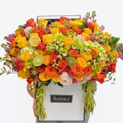 A lavish Florence box overflowing with vibrant orange and yellow roses, green orchids, and fresh citrus fruit slices.