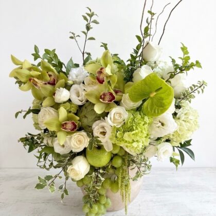 An elegant arrangement of white roses, green orchids, and anthurium accented with fresh grapes in a stone vase.