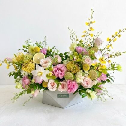 An elegant floral arrangement with pink roses, yellow pincushion proteas, and fresh lime slices in a grey geometric vase.