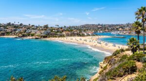 Scenic view of Corona del Mar beach with turquoise water, sandy shores, and luxury homes under a clear blue sky.