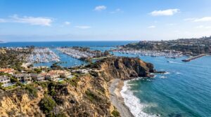 Aerial view of Dana Point Harbor with luxury homes, rocky cliffs, and a marina filled with boats under a blue sky.